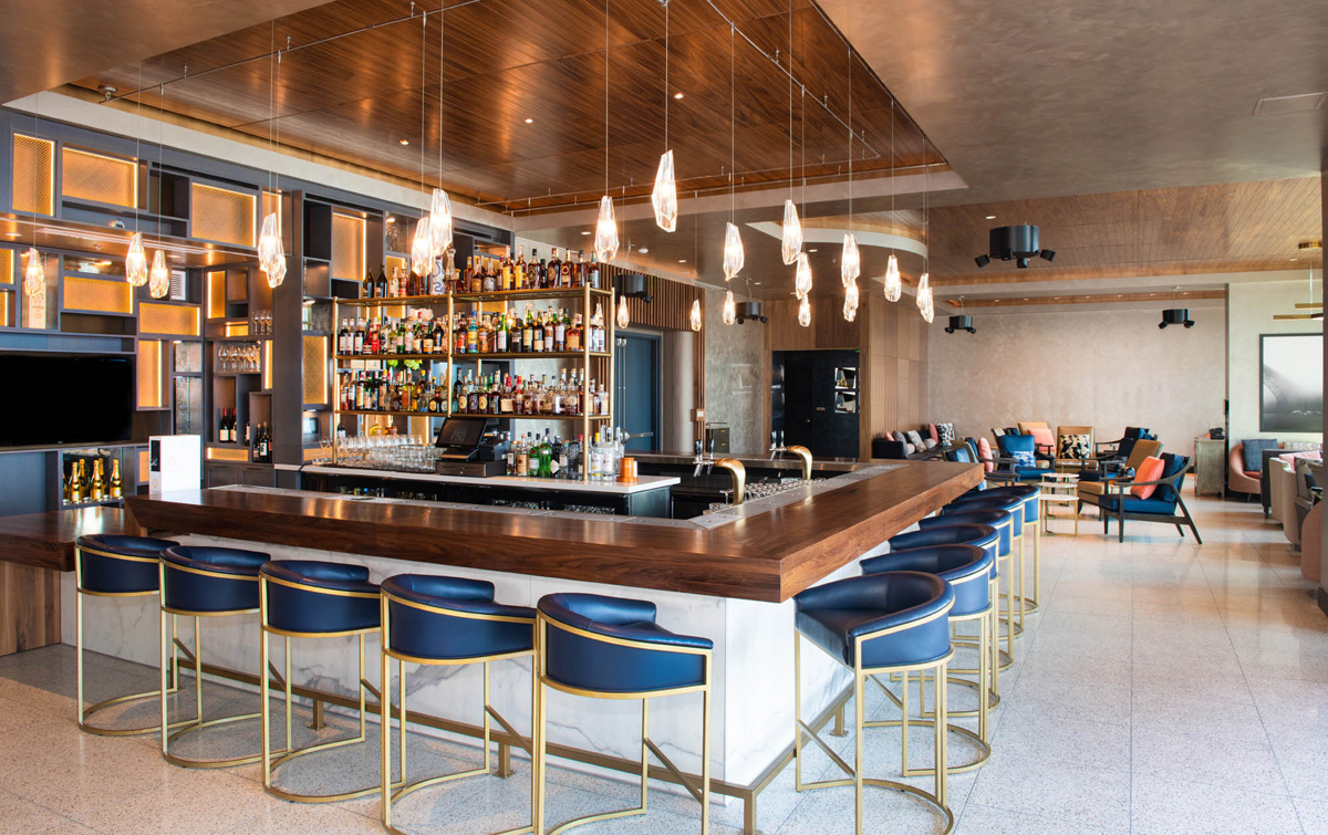 A wide view of a bar with a long counter, blue leather stools, shelves of bottles, and a row of hanging lights.